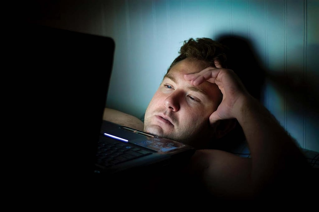a man lies in a dark room staring at a computer screen, he looks very tired