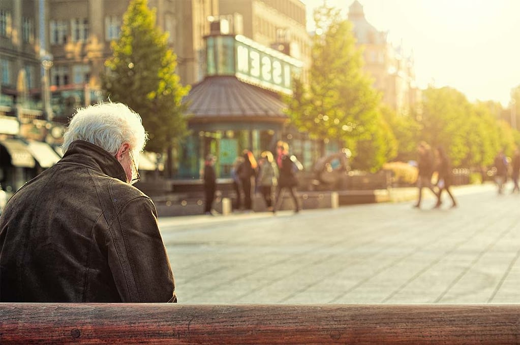 an elderly man sits on a bench in a city square, looking down