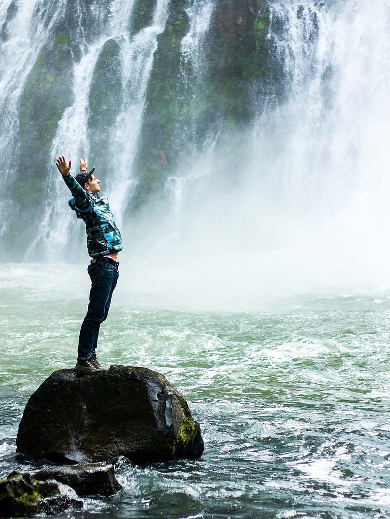 a young man stands on a rock in front of a waterfall raising his arms